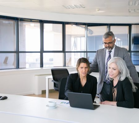 Business team of three watching presentation on pc monitor, discussing project, sitting at workplace and pointing at display. Copy space. Business meeting concept
